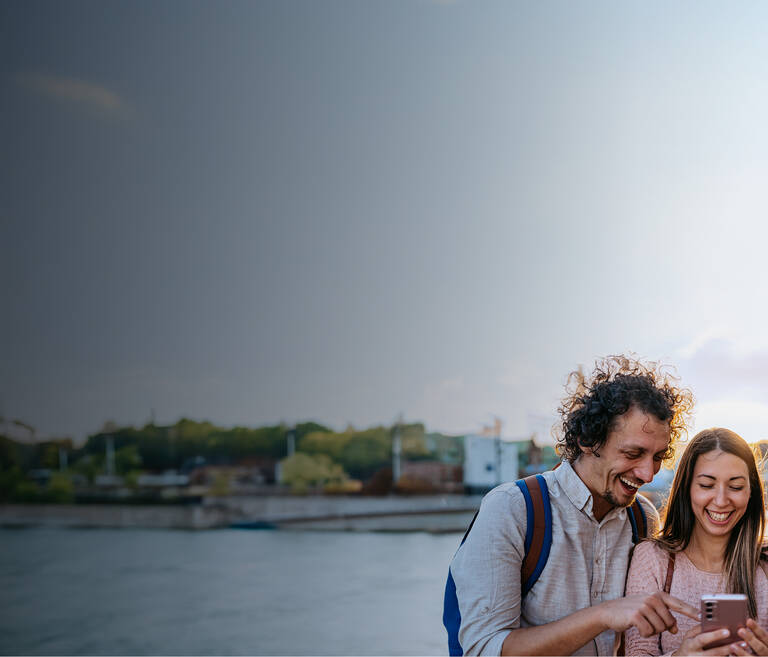 Drei junge Freunde, ein Mann und 2 Frauen, sind in einer Stadt und schauen zusammen lachend auf ein Smartphone.