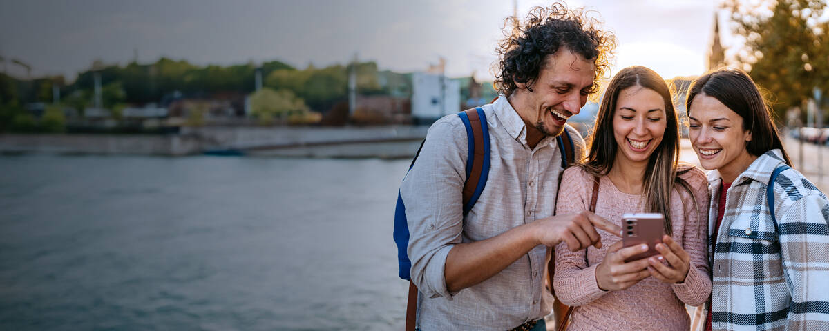 Drei junge Freunde, ein Mann und 2 Frauen, sind in einer Stadt und schauen zusammen lachend auf ein Smartphone.