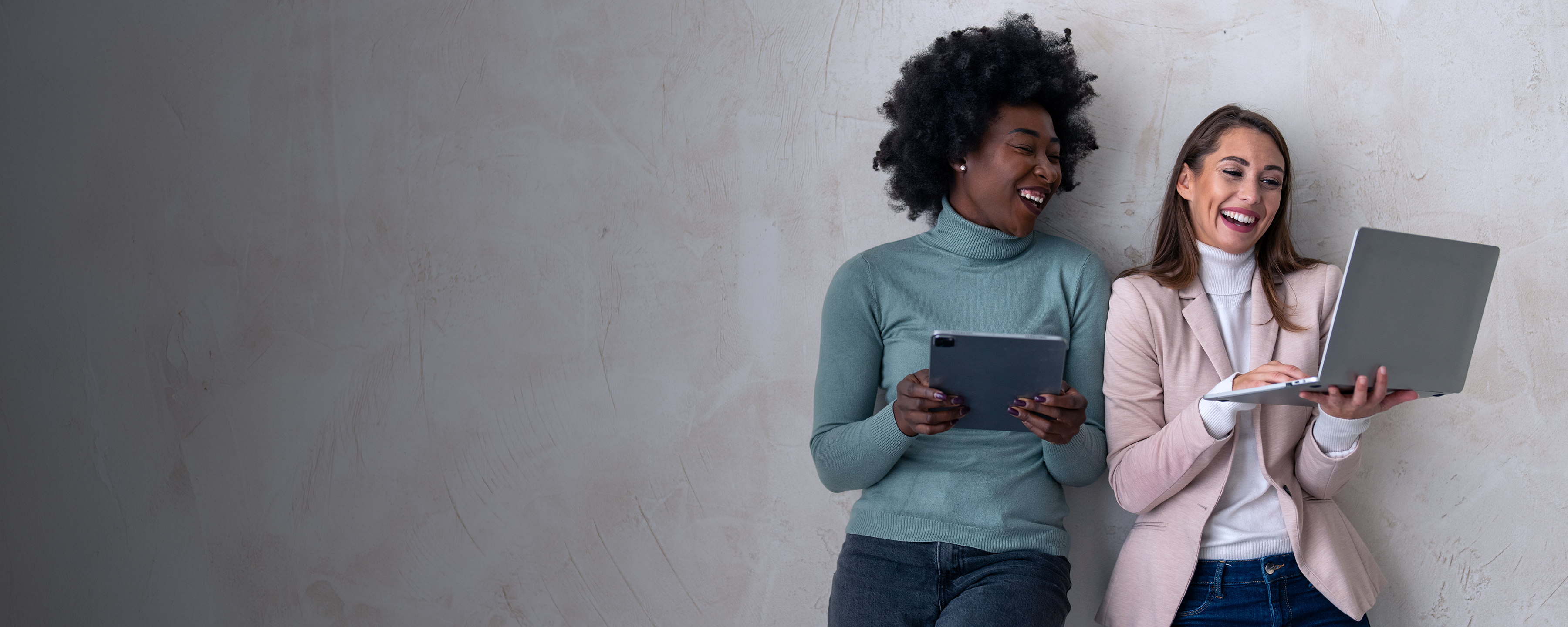 Zwei Frauen stehen nebeneinander an einer Wand angelehnt. Die linke Frau hält ein Tablet und die rechte Frau hält einen Laptop in der Hand