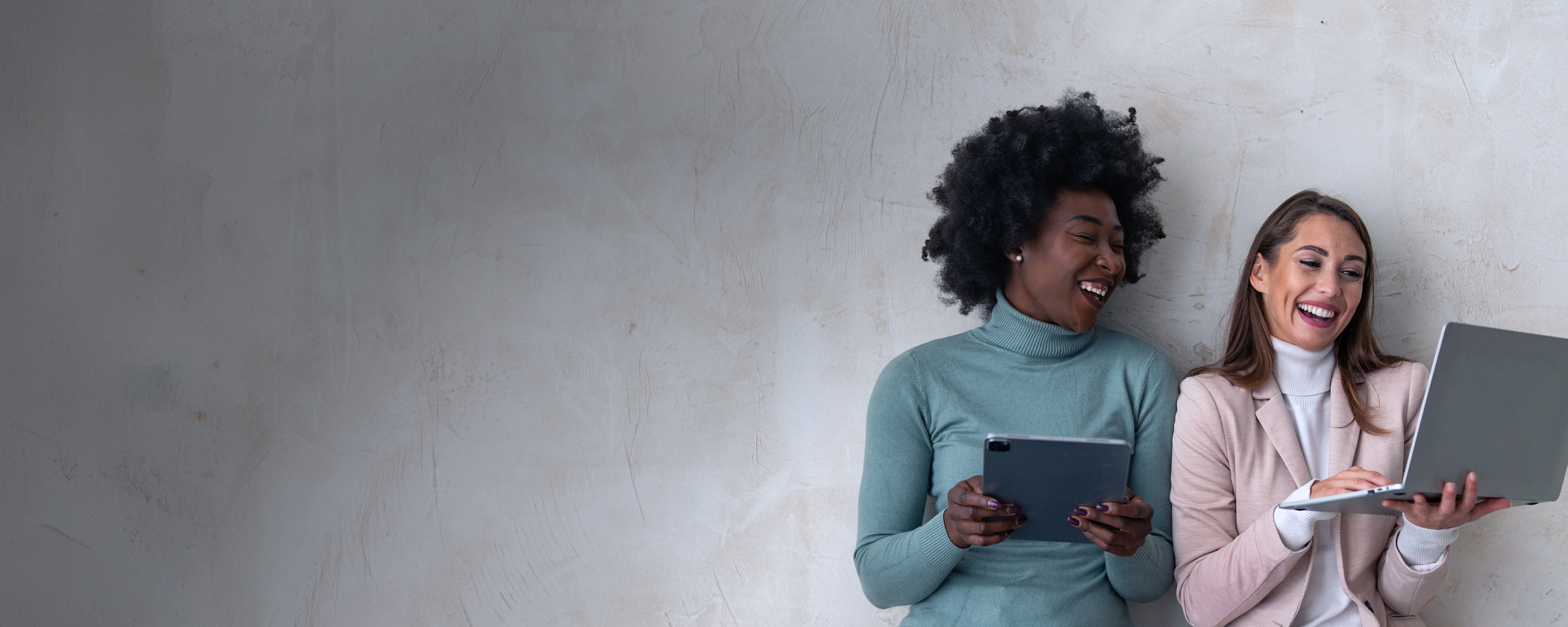 Zwei Frauen stehen nebeneinander an einer Wand angelehnt. Die linke Frau hält ein Tablet und die rechte Frau hält einen Laptop in der Hand
