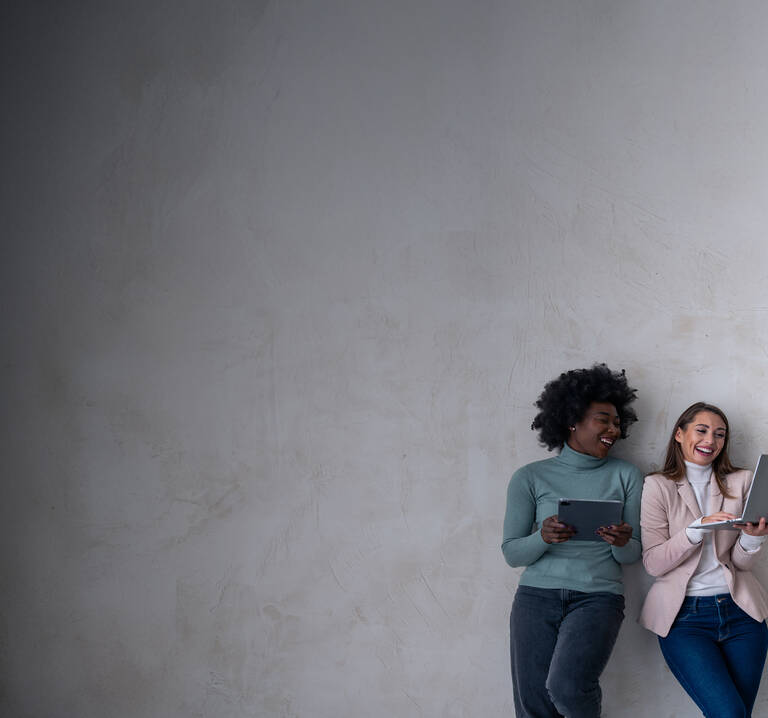 Zwei Frauen stehen nebeneinander an einer Wand angelehnt. Die linke Frau hält ein Tablet und die rechte Frau hält einen Laptop in der Hand